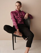 Model seated in the Eloise Plum blouse in satin with black trousers, legs crossed.