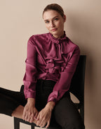 Model seated on a chair in the Eloise Plum blouse in satin with ruffles, worn with black trousers.