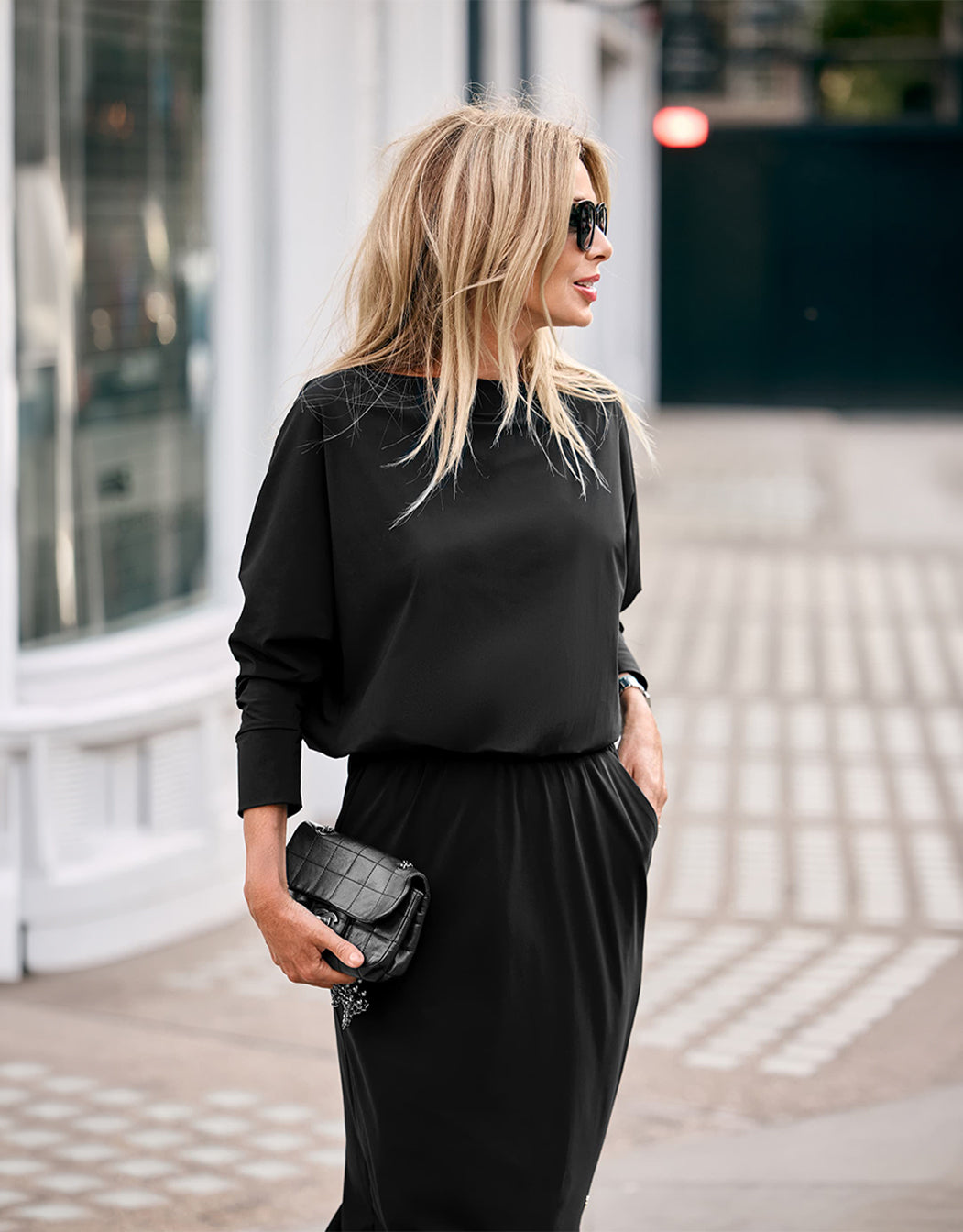 Model staand in de Hillary Black jurk van jersey lycra met een clutch, van opzij gezien.