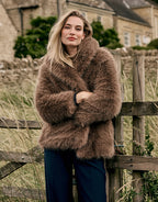 Model steht an einem Holzzaun im Isolde Dark Taupe Mantel aus Kunstfell mit marineblauer Hose.