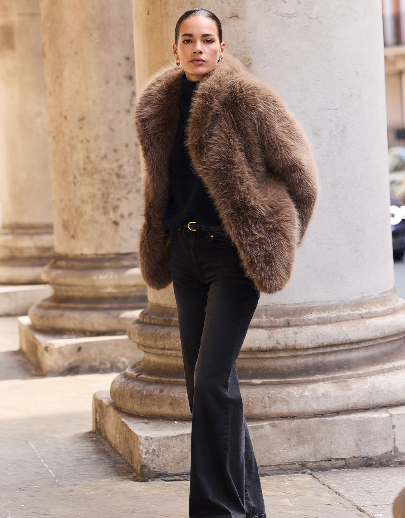 Model steht an Steinpfeilern im Isolde Dark Taupe Mantel aus Kunstfell mit dunkler ausgestellter Jeans.