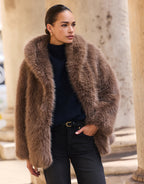 Model im Isolde Dark Taupe Mantel aus Kunstfell über einem marineblauen Pullover, mit den Händen in den Taschen.