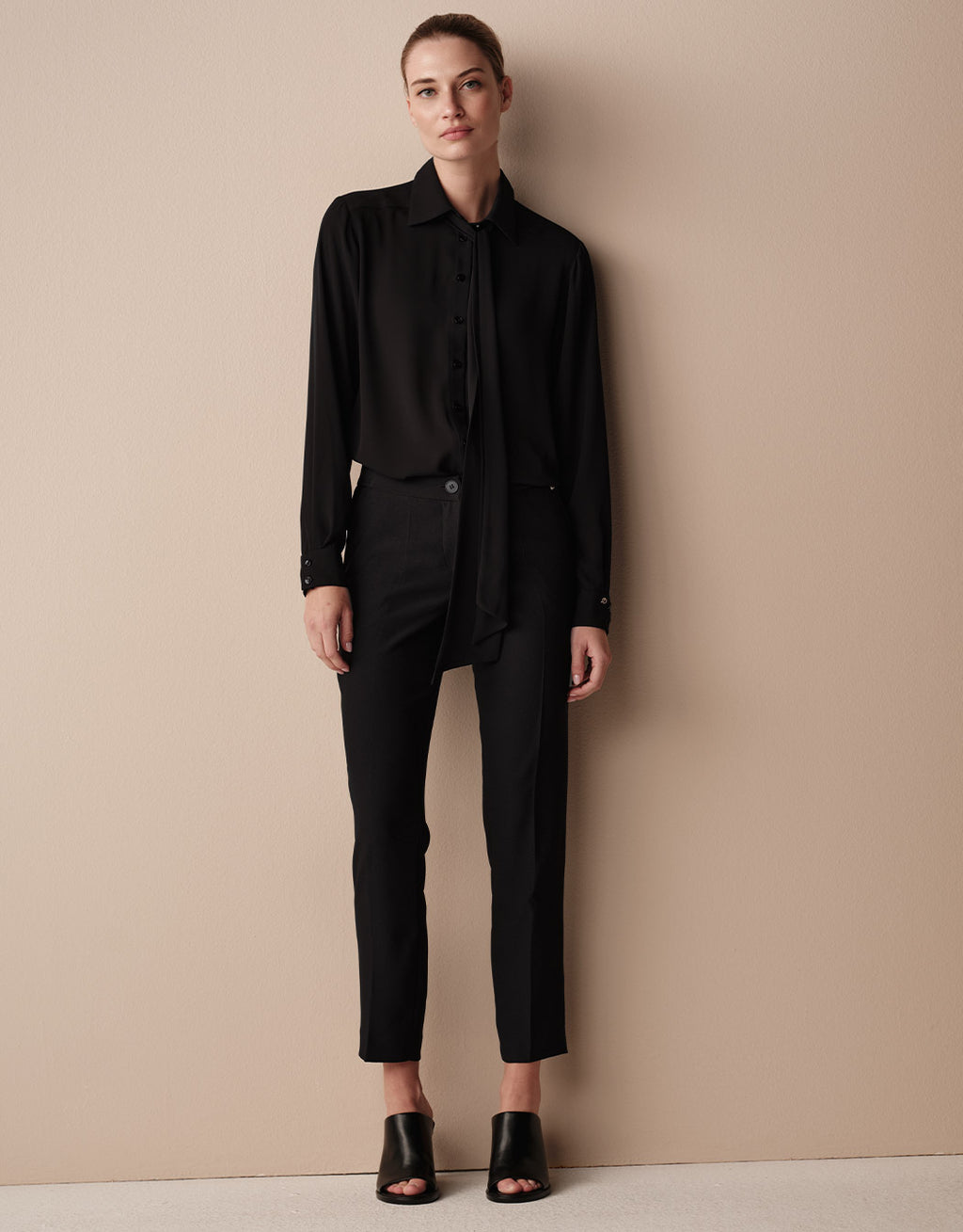 Model steht in der Kris Black Bluse aus Crêpe Georgette mit offener Schleife und einer schwarzen Hose.