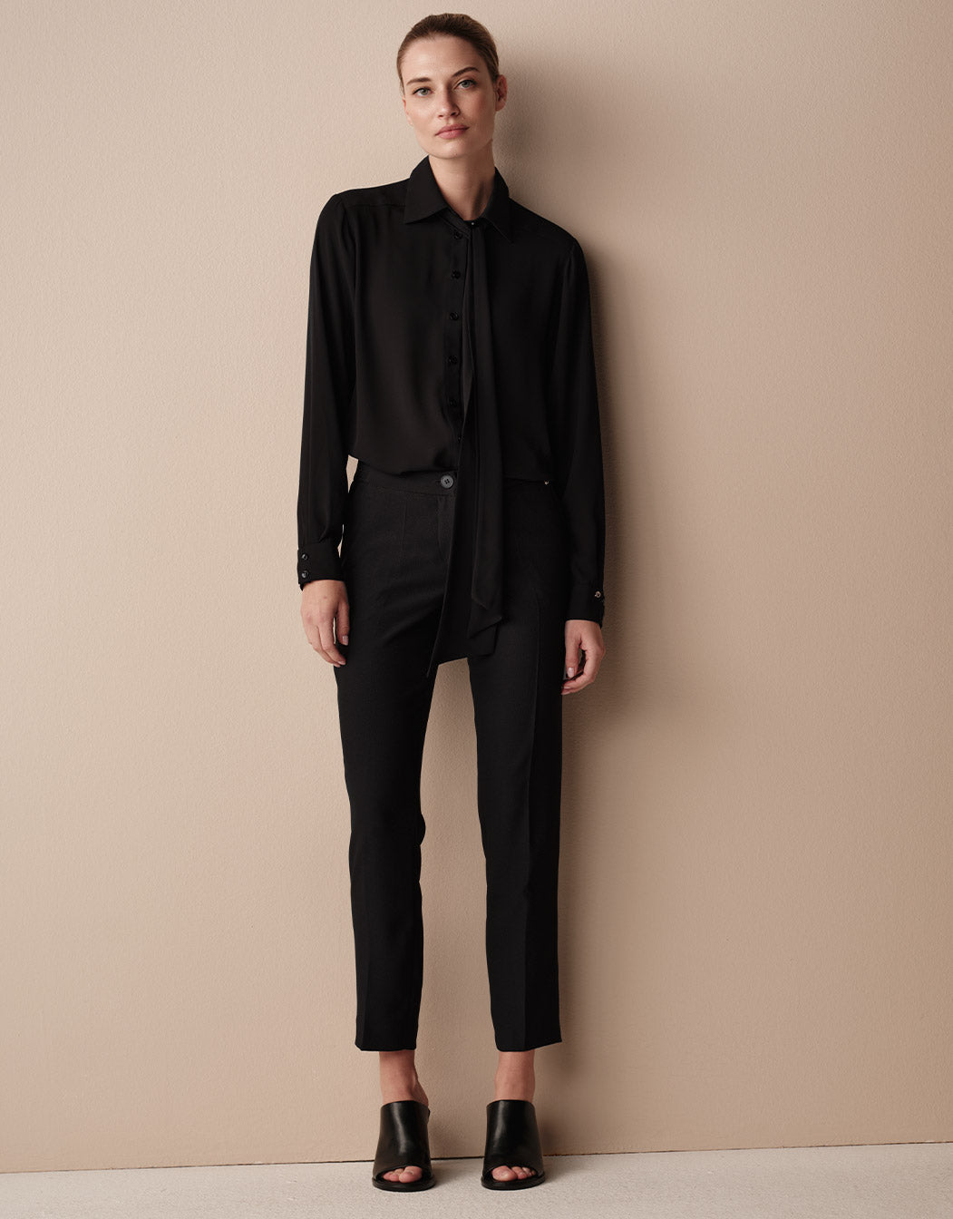 Model steht in der Kris Black Bluse aus Crêpe Georgette mit offener Schleife und einer schwarzen Hose.