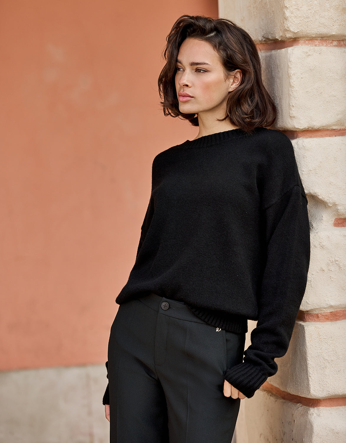 Model leaning in the Lucienne Black jumper in viscose knit with black trousers.