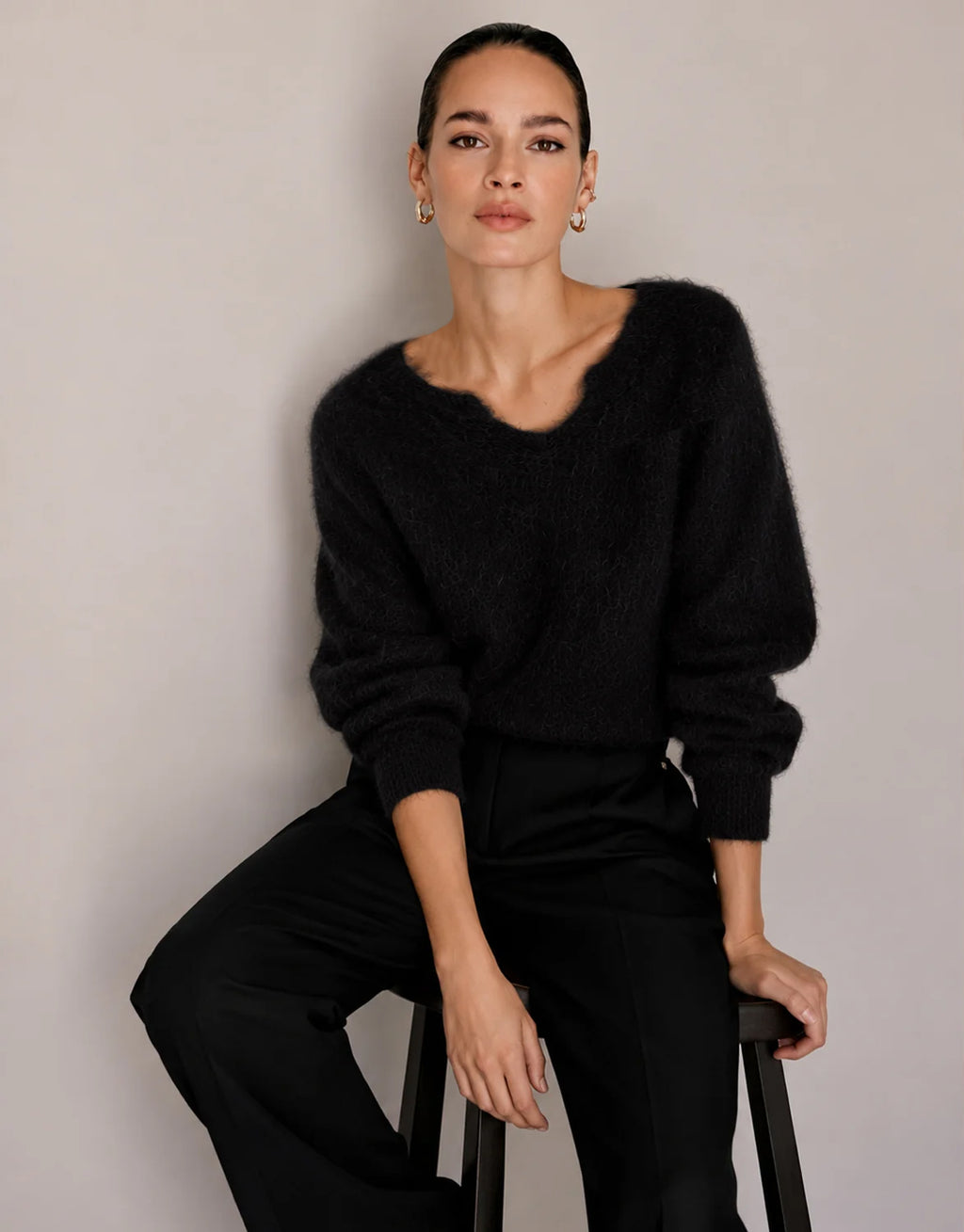 Model zittend op een kruk in de Stefania Black trui van alpacamix met een zwarte broek.
