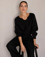 Model zittend op een kruk in de Stefania Black trui van alpacamix met een zwarte broek.
