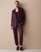 Model standing against a beige wall in the Juliette Deep Aubergine Satin blazer and Verona trousers.