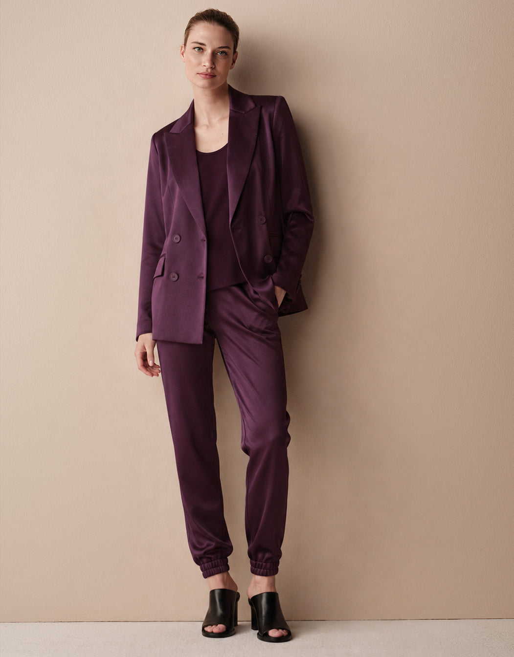 Model staat tegen een beige muur in de Juliette Deep Aubergine Satin blazer en Verona broek.