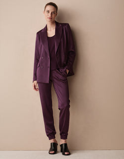 Model standing against a beige wall in the Juliette Deep Aubergine Satin blazer and Verona trousers.