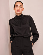 Model steht in der Vittoria Black Bluse aus Satin mit hohem Kragen, mit den Händen in den Taschen.