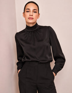 Model steht in der Vittoria Black Bluse aus Satin mit hohem Kragen, mit den Händen in den Taschen.