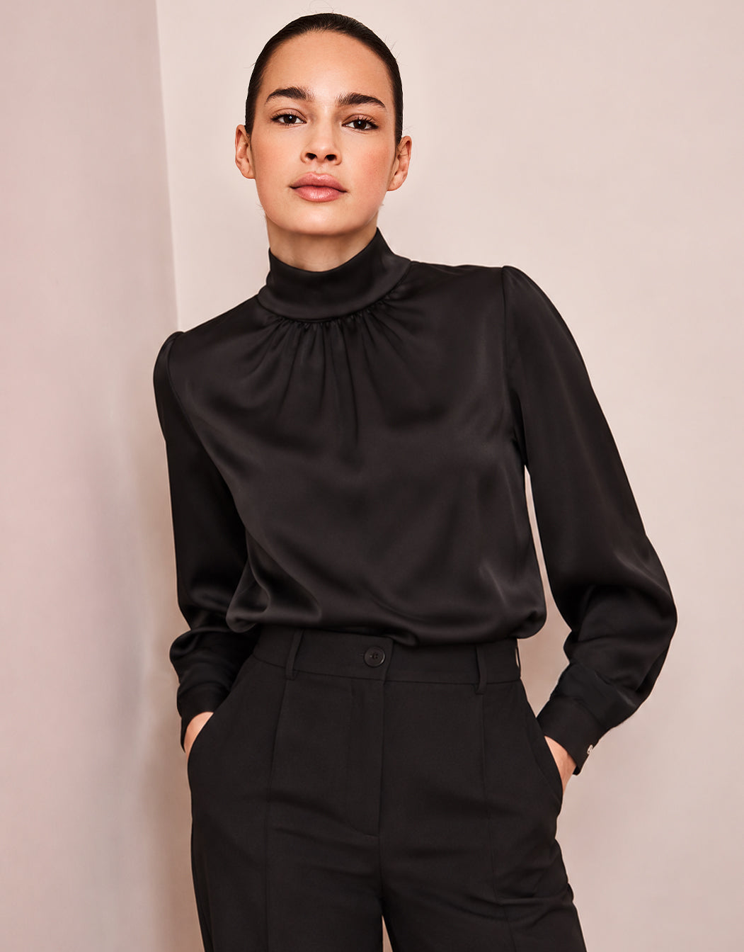 Model steht in der Vittoria Black Bluse aus Satin mit hohem Kragen, mit den Händen in den Taschen.