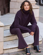 Model seated on steps in the Giovanna Deep Aubergine jumper in alpaca blend with matching trousers.