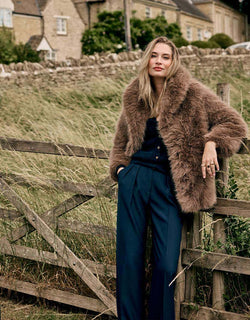 Model standing in the Isolde Dark Taupe coat in faux fur with wide navy trousers.