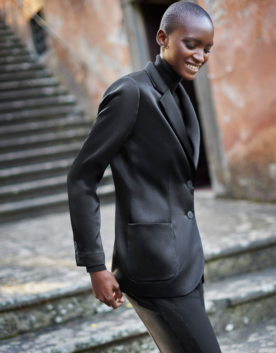 Model standing in the Carey Black Satin blazer in crepe satin over a matching top and trousers.
