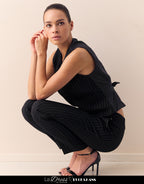 Woman dressed in an elegant black pinstripe pantsuit with a sleeveless top and fashionable sandals against a minimalist background.