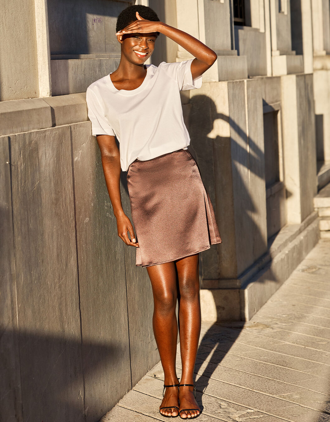 Model leaning in the Gigi Cacao Brown Satin skirt in crepe satin with a white T-shirt.