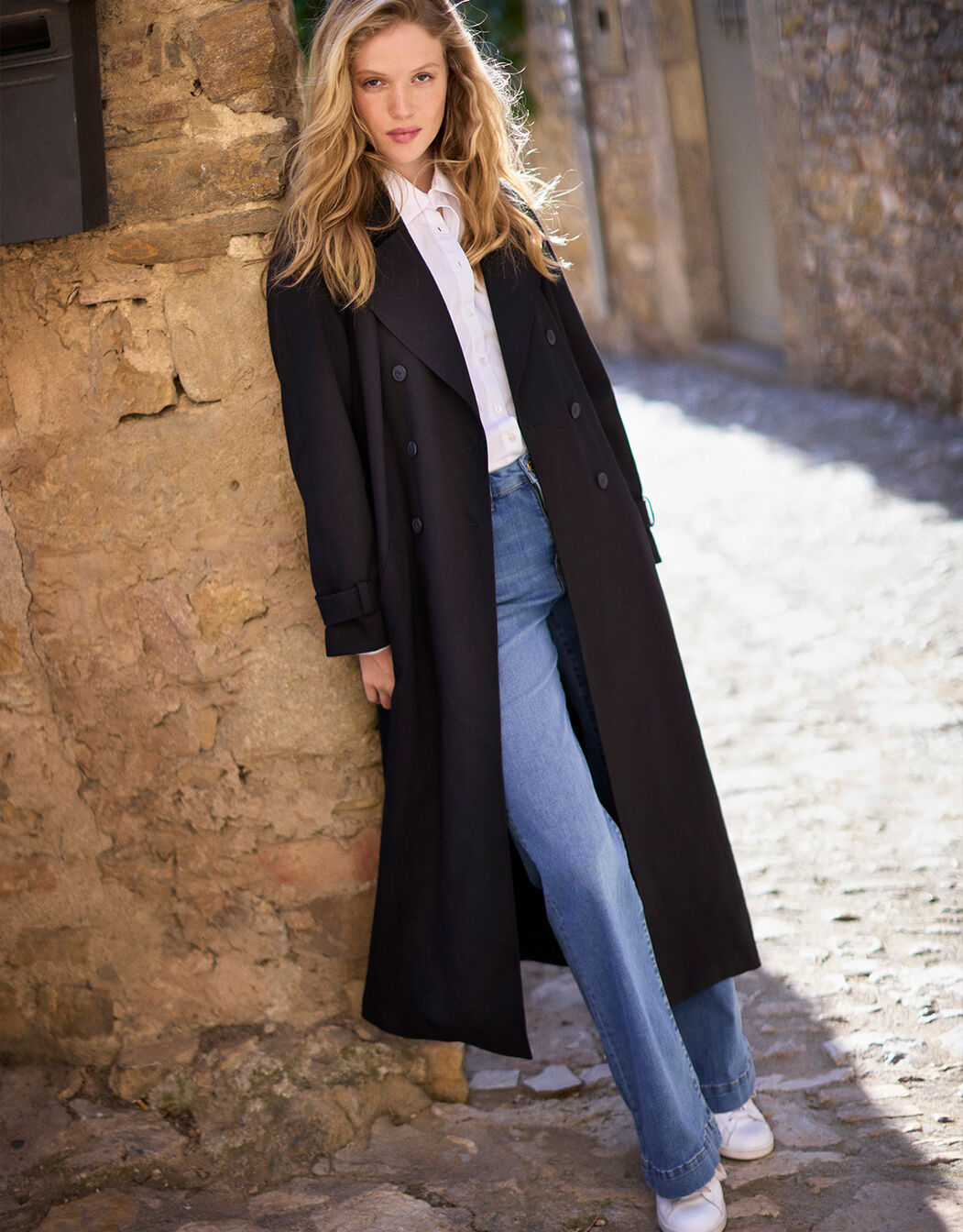 Model in the long Alina Black trench coat worn open over a white shirt and jeans.