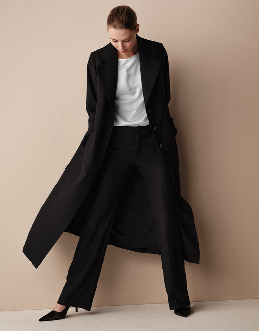 Model walking in the Vivianne Black coat in crepe satin, a long single-breasted style.