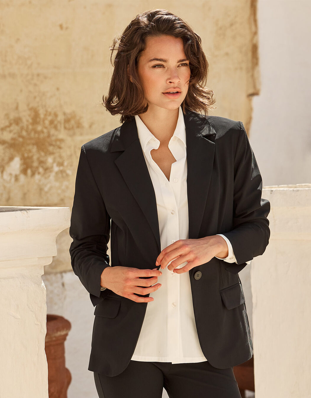 Woman in black blazer and white blouse stands against a beige stone wall, exuding elegance and professionalism.